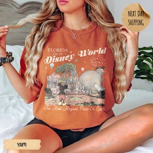May include: Orange t-shirt with a graphic of Florida Disney World, featuring Mickey Mouse, a large sphere, and the text "The Most Magical Place On Earth." The shirt has a vintage style.