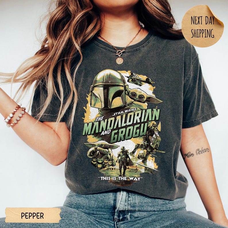 Pu&ograve; includere: T-shirt grigio scuro con una grafica della serie The Mandalorian. Il design include il casco Mandaloriano, Grogu e il testo "The Mandalorian and Grogu". La frase "This is the Way" &egrave; stampata anche sulla maglietta.
