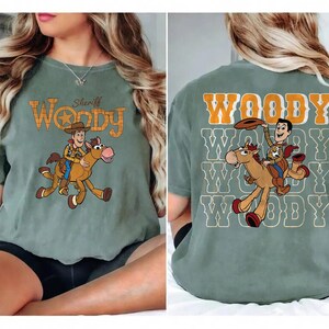 May include: Sage green t-shirt featuring a cartoon illustration of Woody riding a horse. The front of the shirt has the text "Sheriff Woody" in orange and brown. The back of the shirt has the word "Woody" repeated multiple times.