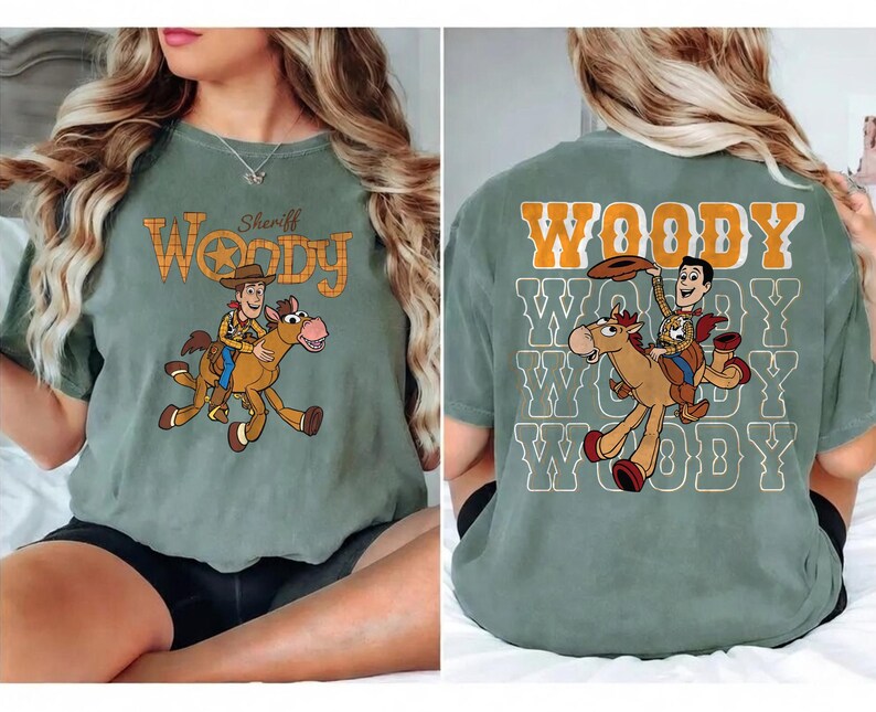 May include: Sage green t-shirt featuring a cartoon illustration of Woody riding a horse. The front of the shirt has the text "Sheriff Woody" in orange and brown. The back of the shirt has the word "Woody" repeated multiple times.