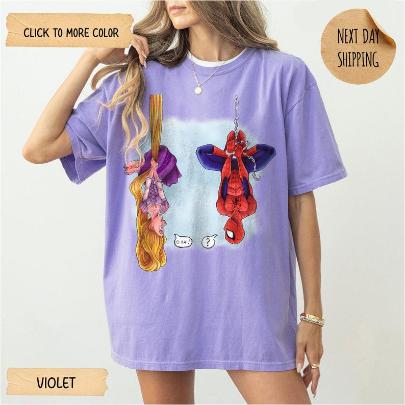 Op de afbeelding: Een violetkleurig T-shirt met korte mouwen en een grafische print van een langharig personage dat aan haar haar hangt en een rood-blauwe superheld die aan een web hangt. Het personage links heeft een tekstballon met O HAI! en de superheld een vraagteken.