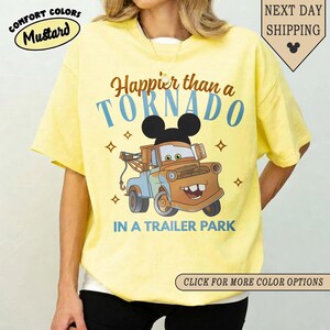 K&ouml;nnte beinhalten: Senfgelbes T-Shirt mit einem Cartoon-Abschleppwagen-Design. Das Design enth&auml;lt den Text "Happier than a TORNADO IN A TRAILER PARK" und einen Mickey Mouse-Kopf. Das Shirt tr&auml;gt auch die Aufschrift "COMFORT COLORS" und "NEXT DAY SHIPPING".