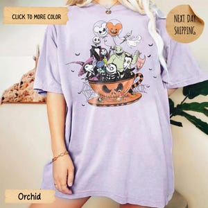 K&ouml;nnte beinhalten: Orchideenfarbenes T-Shirt mit einem Halloween-Motiv. Das Design zeigt Charaktere aus "Nightmare Before Christmas" in einer Jack-o'-Lantern-Tasse. Das Shirt hat kurze &Auml;rmel und eine lockere Passform. Der Text "Click to More Color" und "Next Day Shipping" sind sichtbar.