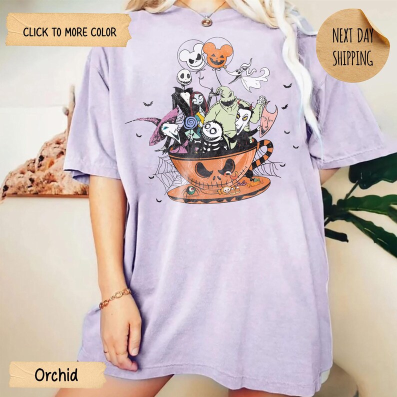 K&ouml;nnte beinhalten: Orchideenfarbenes T-Shirt mit einem Halloween-Motiv. Das Design zeigt Charaktere aus "Nightmare Before Christmas" in einer Jack-o'-Lantern-Tasse. Das Shirt hat kurze &Auml;rmel und eine lockere Passform. Der Text "Click to More Color" und "Next Day Shipping" sind sichtbar.