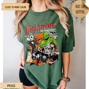 May include: A moss green short-sleeve t-shirt featuring a graphic print with the text Nightmare on Main Street. The design includes characters from The Nightmare Before Christmas, a large orange moon, a tombstone, a pumpkin, and spider webs.