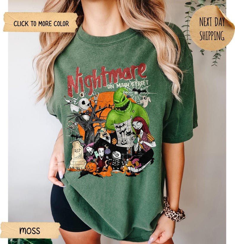 May include: A moss green short-sleeve t-shirt featuring a graphic print with the text Nightmare on Main Street. The design includes characters from The Nightmare Before Christmas, a large orange moon, a tombstone, a pumpkin, and spider webs.