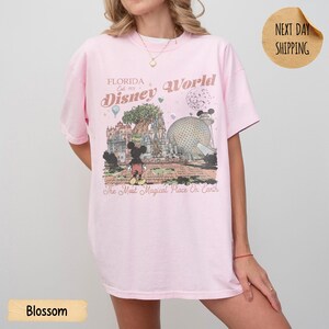 May include: A light pink t-shirt featuring a graphic of Disney World in Florida. The design includes Mickey Mouse, a castle, and the text "Florida Disney World" and "The Most Magical Place On Earth". The shirt has short sleeves.