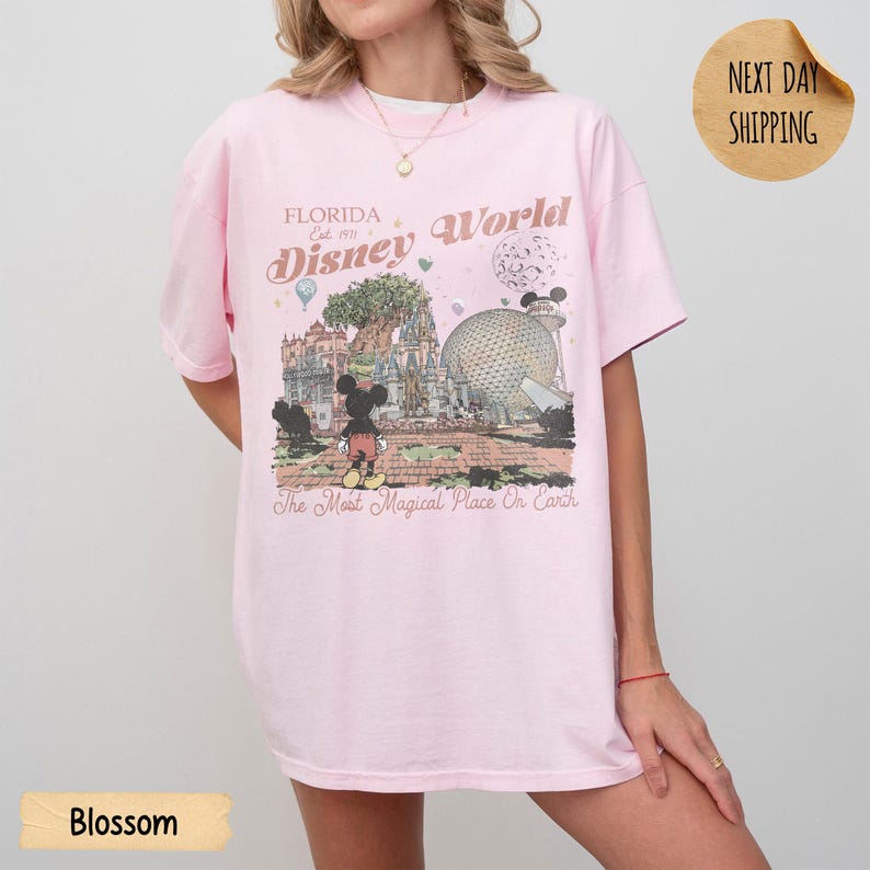 May include: A light pink t-shirt featuring a graphic of Disney World in Florida. The design includes Mickey Mouse, a castle, and the text "Florida Disney World" and "The Most Magical Place On Earth". The shirt has short sleeves.