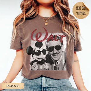May include: Espresso-colored t-shirt featuring a graphic of Mickey Mouse and Walt Disney, with the word "Walt" in red script. Mickey is wearing sunglasses. The image is in a vintage style.