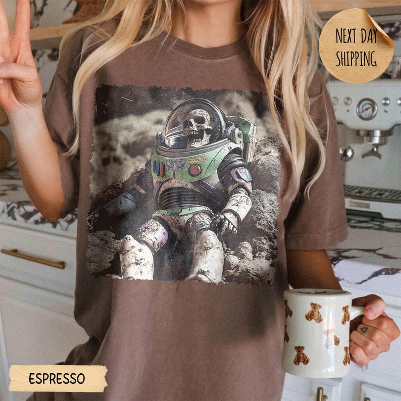 May include: A brown t-shirt featuring a graphic print of a skeleton wearing a space suit helmet and armor. The shirt is paired with a white mug decorated with a repeating pattern of small brown teddy bears. The text ESPRESSO is visible below.