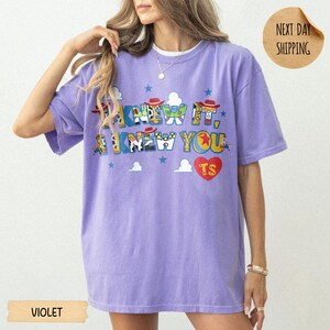 May include: A violet short-sleeve t-shirt featuring the text I KNEW IT, I KNEW YOU in a multi-colored graphic font with character-inspired patterns. A red heart with the letters TS is printed below the text, accompanied by small blue stars.