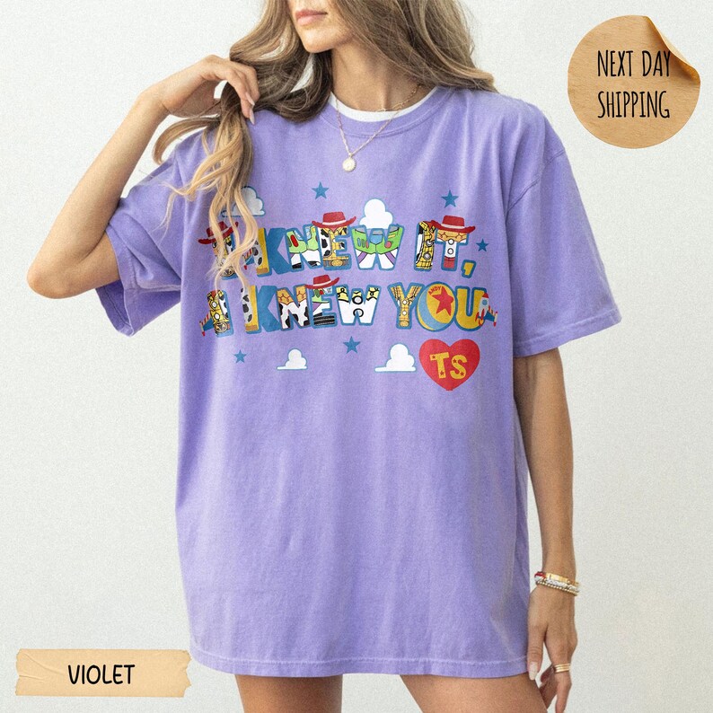 May include: A violet short-sleeve t-shirt featuring the text I KNEW IT, I KNEW YOU in a multi-colored graphic font with character-inspired patterns. A red heart with the letters TS is printed below the text, accompanied by small blue stars.