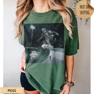 May include: Moss green t-shirt featuring a graphic of a character skateboarding, with a smaller character on a skateboard below. The image has a distressed, vintage look. The shirt is a relaxed fit.