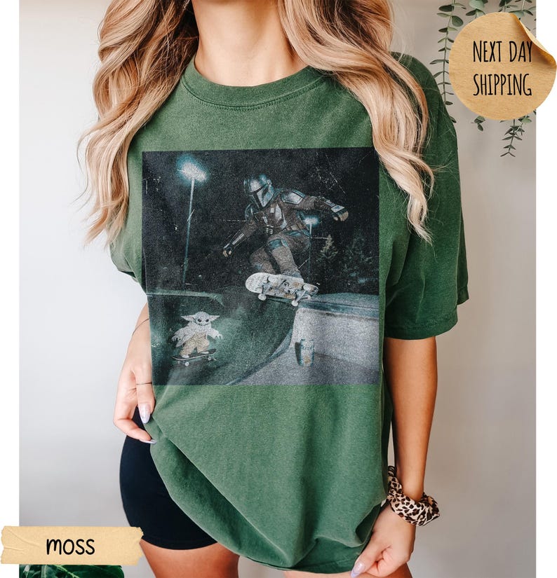 May include: Moss green t-shirt featuring a graphic of a character skateboarding, with a smaller character on a skateboard below. The image has a distressed, vintage look. The shirt is a relaxed fit.