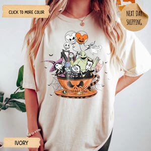 K&ouml;nnte beinhalten: Elfenbeinfarbenes T-Shirt mit einem Halloween-Motiv. Die Grafik zeigt Charaktere aus Nightmare Before Christmas in einer Jack-o'-Lantern-Tasse, mit Luftballons und Spinnweben. Das T-Shirt hat einen Rundhalsausschnitt und kurze &Auml;rmel.