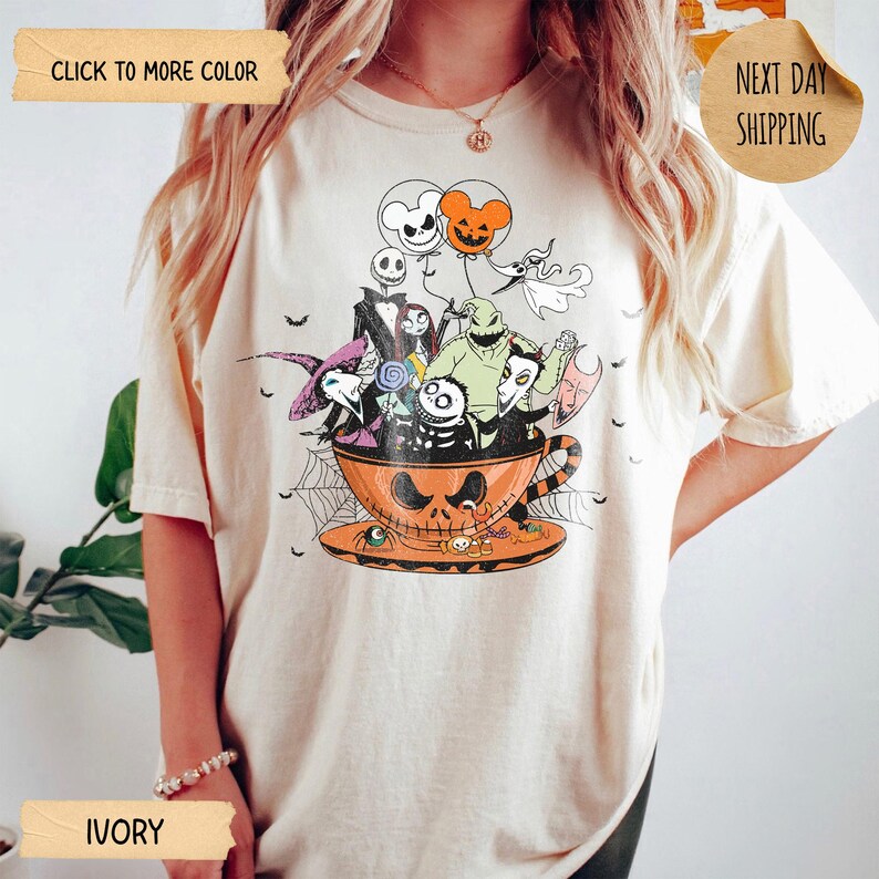 K&ouml;nnte beinhalten: Elfenbeinfarbenes T-Shirt mit einem Halloween-Motiv. Die Grafik zeigt Charaktere aus Nightmare Before Christmas in einer Jack-o'-Lantern-Tasse, mit Luftballons und Spinnweben. Das T-Shirt hat einen Rundhalsausschnitt und kurze &Auml;rmel.