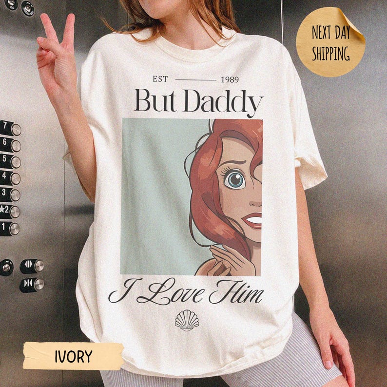 Op de afbeelding: Een ivoorkleurig t-shirt met een grafische print van een roodharig personage met blauwe ogen. De tekst luidt EST 1989, But Daddy, en I Love Him, vergezeld door een klein schelppictogram. Het ontwerp is gecentreerd op de voorkant van het shirt.