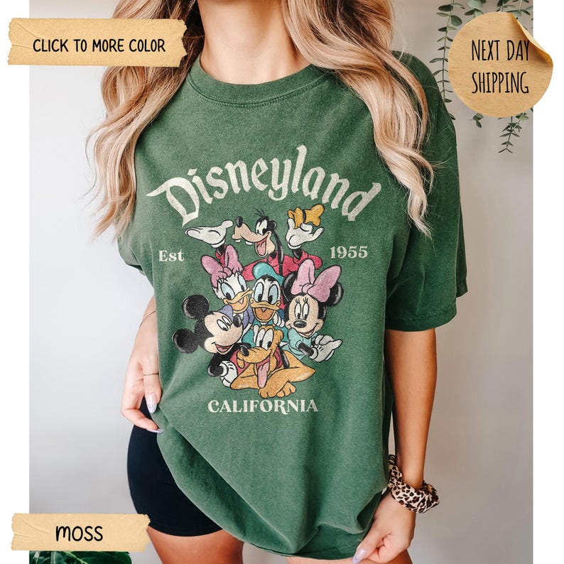 Op de afbeelding: Mosgroen T-shirt met een Disneyland-afbeelding met Mickey Mouse en vrienden. De tekst "Disneyland Est 1955 California" staat op de voorkant. Het shirt heeft korte mouwen.