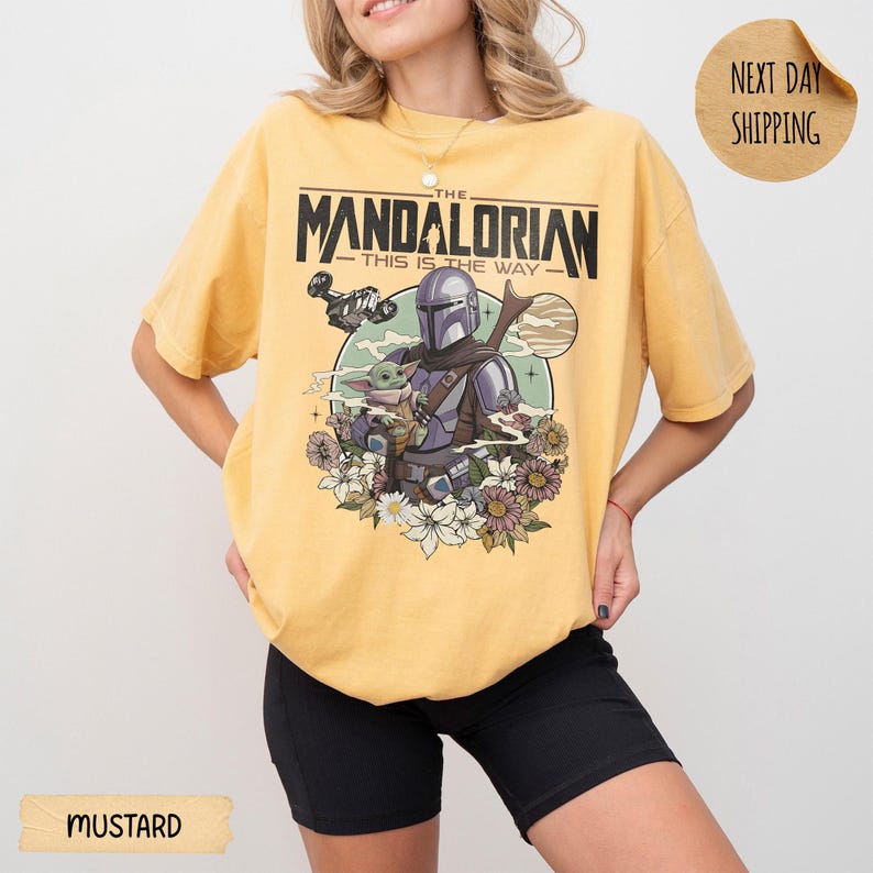 May include: Mustard-colored t-shirt featuring a graphic of The Mandalorian and Grogu surrounded by flowers. The text "THE MANDALORIAN - THIS IS THE WAY" is printed above the image. The shirt has short sleeves.