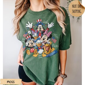 May include: A moss green short-sleeve t-shirt featuring a graphic print of Disney characters including Mickey Mouse, Donald Duck, Goofy, Minnie Mouse, Daisy Duck, and Pluto wearing patriotic hats. The text reads America 250th Anniversary 1776-2026.