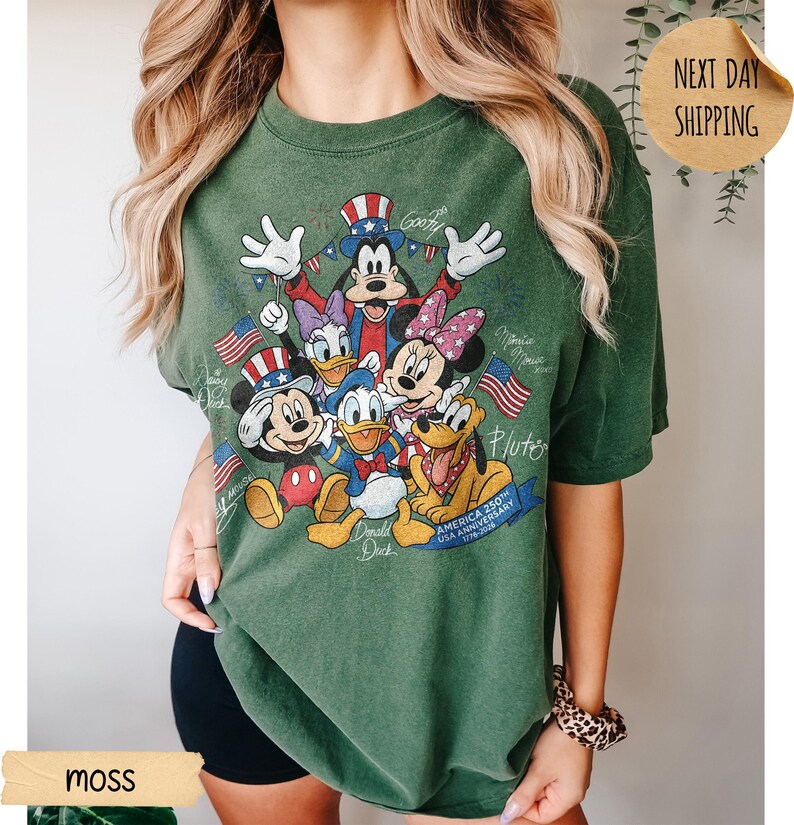 May include: A moss green short-sleeve t-shirt featuring a graphic print of Disney characters including Mickey Mouse, Donald Duck, Goofy, Minnie Mouse, Daisy Duck, and Pluto wearing patriotic hats. The text reads America 250th Anniversary 1776-2026.