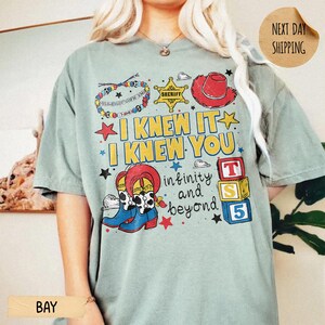 May include: A sage green short-sleeve t-shirt featuring a colorful graphic print with a beaded bracelet, a yellow sheriff star, a red cowboy hat, and blue cowboy boots. The text reads I KNEW IT I KNEW YOU infinity and beyond with alphabet blocks.