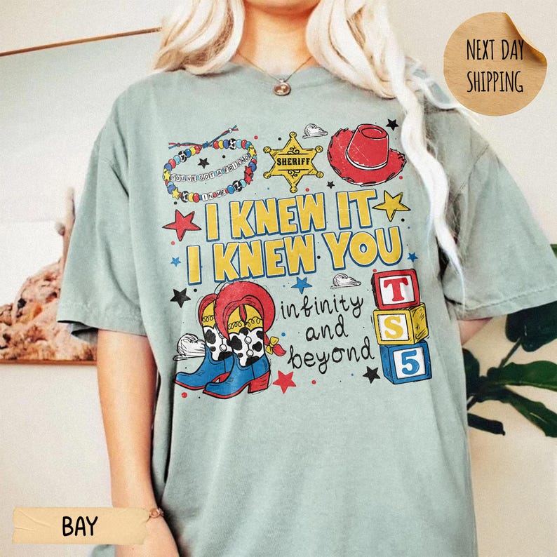 May include: A sage green short-sleeve t-shirt featuring a colorful graphic print with a beaded bracelet, a yellow sheriff star, a red cowboy hat, and blue cowboy boots. The text reads I KNEW IT I KNEW YOU infinity and beyond with alphabet blocks.