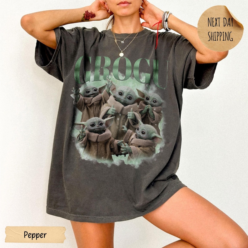 May include: A charcoal gray short-sleeve t-shirt featuring a large graphic print of five green alien creatures in brown robes. The text GROGU is printed in large green letters above the characters. The shirt has a loose, relaxed fit design.