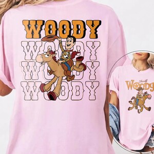 May include: Light pink t-shirt featuring a cartoon graphic of Woody from Toy Story riding a horse. The word "WOODY" is printed in orange and black above the graphic, with the same word repeated in a white outline below.