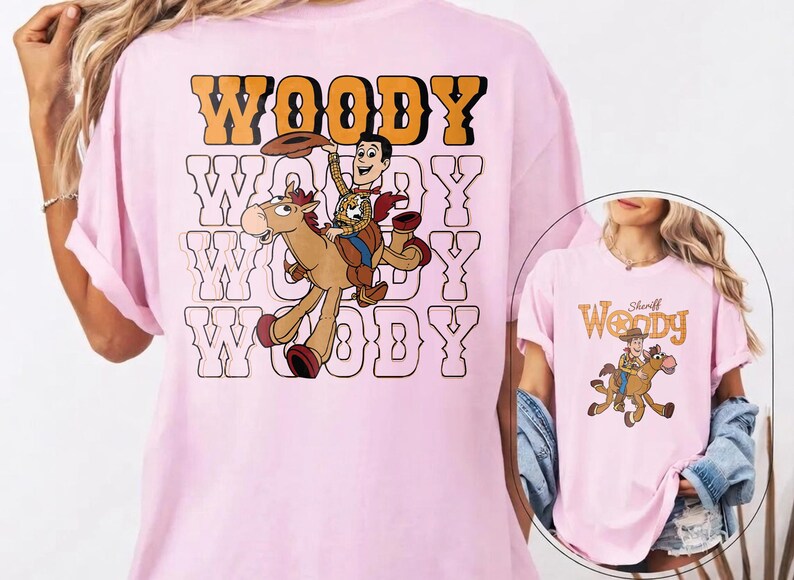 May include: Light pink t-shirt featuring a cartoon graphic of Woody from Toy Story riding a horse. The word "WOODY" is printed in orange and black above the graphic, with the same word repeated in a white outline below.