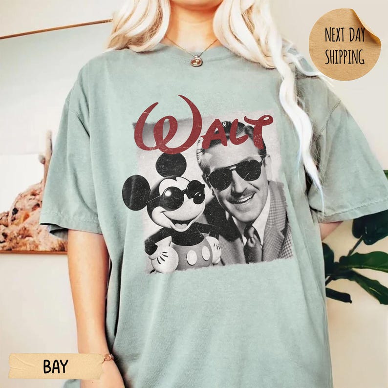 May include: A light green t-shirt featuring a vintage graphic of Mickey Mouse and Walt Disney, both wearing sunglasses. The word "Walt" is printed above the image in a distressed red font. The shirt has short sleeves.