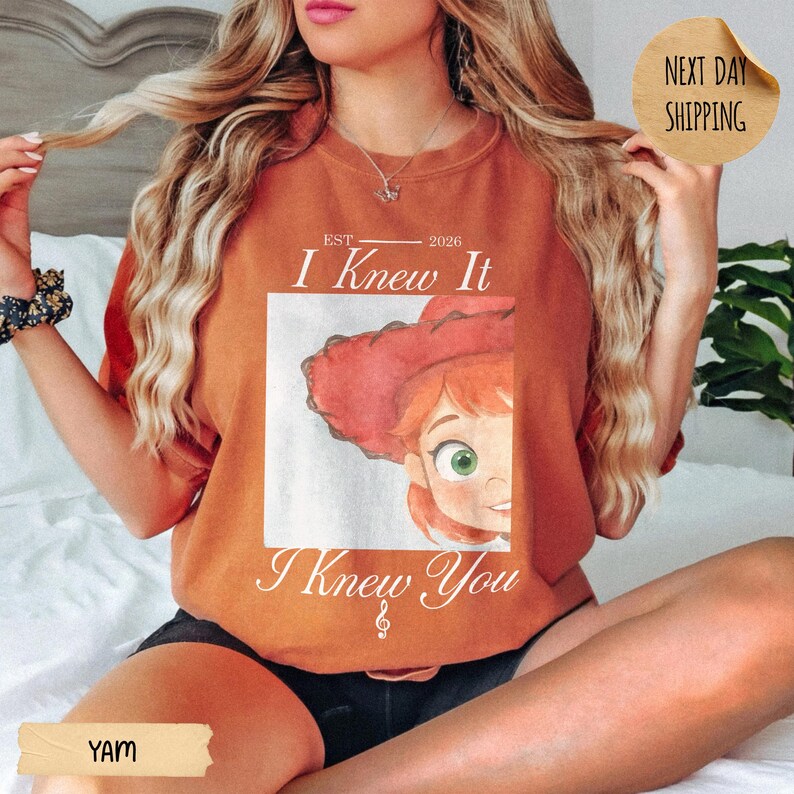 May include: A rust-colored crewneck t-shirt featuring a graphic of a red-haired character in a red cowboy hat. The text reads "EST 2026 I Knew It I Knew You" with a small musical note symbol. The shirt has short sleeves and a round neckline.