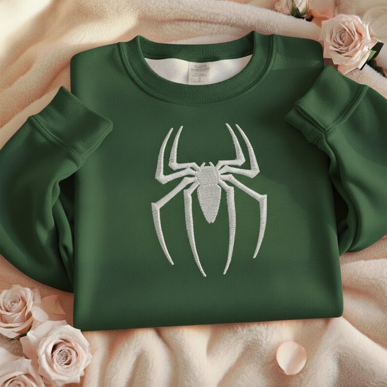 Puede incluir: Una sudadera verde oscuro con un dise&ntilde;o de ara&ntilde;a blanco bordado en la parte delantera. La sudadera tiene cuello redondo y mangas largas con pu&ntilde;os acanalados. La imagen est&aacute; sobre un fondo suave y claro con rosas.