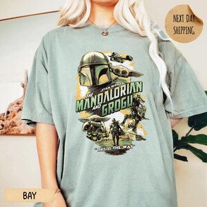 Pu&ograve; includere: Una maglietta verde chiaro con una grafica della serie Star Wars, "The Mandalorian e Grogu". Il design include il casco Mandaloriano, Grogu e la frase "This is the Way".