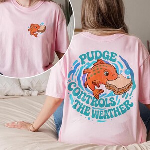 May include: A light pink t-shirt with a cartoon orange fish and sandwich design. The back of the shirt has the text "Pudge Controls the Weather". The front has a smaller version of the fish and sandwich graphic.