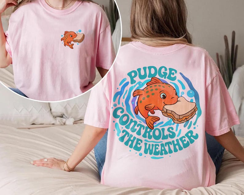 May include: A light pink t-shirt with a cartoon orange fish and sandwich design. The back of the shirt has the text "Pudge Controls the Weather". The front has a smaller version of the fish and sandwich graphic.