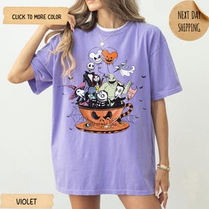 K&ouml;nnte beinhalten: Violettes T-Shirt mit einem Halloween-Motiv. Das Design zeigt Charaktere aus "Nightmare Before Christmas" in einer Jack-o'-Lantern-Tasse. Das Shirt hat eine lockere Passform und kurze &Auml;rmel. Der Text "CLICK TO MORE COLOR" und "VIOLET" sind sichtbar.