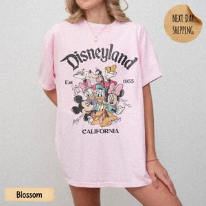 May include: A light pink short-sleeve t-shirt featuring a graphic print of Disney characters including Mickey Mouse, Minnie Mouse, Donald Duck, Daisy Duck, Goofy, and Pluto. The text reads Disneyland, Est 1955, and California in black font.