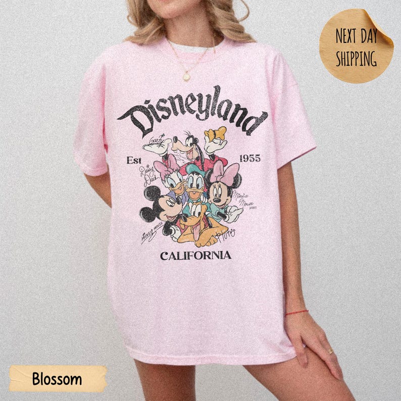 May include: A light pink short-sleeve t-shirt featuring a graphic print of Disney characters including Mickey Mouse, Minnie Mouse, Donald Duck, Daisy Duck, Goofy, and Pluto. The text reads Disneyland, Est 1955, and California in black font.