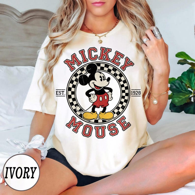 Puede incluir: Camiseta color crema con un gr&aacute;fico de Mickey Mouse. El dise&ntilde;o incluye las palabras "Mickey Mouse" en rojo, un borde a cuadros y el a&ntilde;o "1928". Una prenda casual de inspiraci&oacute;n vintage.
