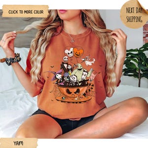 K&ouml;nnte beinhalten: Orangefarbenes T-Shirt mit einem Halloween-Motiv. Das Design zeigt Charaktere aus Nightmare Before Christmas in einer Jack-o'-Lantern-Tasse mit Mickey Mouse-f&ouml;rmigen Luftballons. Das Shirt hat das Wort "YAM" unten.