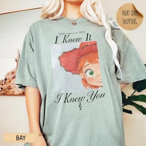 May include: A sage green short-sleeve t-shirt featuring a graphic of a cartoon character with red hair and a red hat. The text above the graphic reads EST. 2026 I Knew It and the text below reads I Knew You with a small musical note symbol.