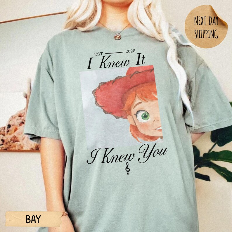 May include: A sage green short-sleeve t-shirt featuring a graphic of a cartoon character with red hair and a red hat. The text above the graphic reads EST. 2026 I Knew It and the text below reads I Knew You with a small musical note symbol.