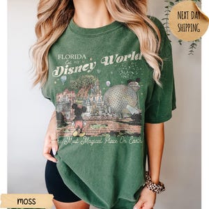 May include: Moss green t-shirt featuring a graphic of Disney World in Florida. The design includes Mickey Mouse, Cinderella's castle, and Spaceship Earth. Text reads "Florida Disney World" and "The Most Magical Place On Earth."