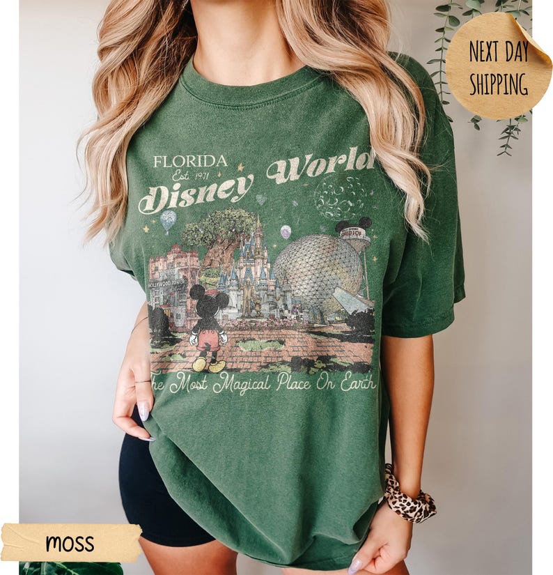 May include: Moss green t-shirt featuring a graphic of Disney World in Florida. The design includes Mickey Mouse, Cinderella's castle, and Spaceship Earth. Text reads "Florida Disney World" and "The Most Magical Place On Earth."