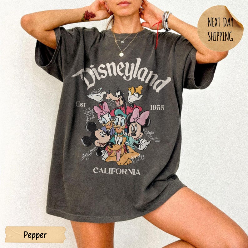 May include: A charcoal gray oversized t-shirt featuring a vintage-style graphic of Disney characters including Mickey Mouse, Minnie Mouse, Donald Duck, Daisy Duck, Goofy, and Pluto. The text reads Disneyland, Est 1955, and California.