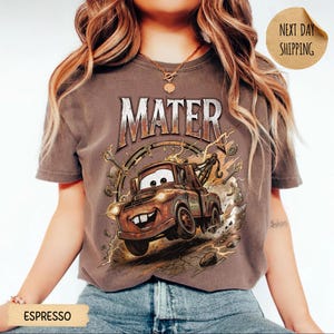 May include: Espresso-coloured t-shirt featuring a cartoon image of Mater from the film Cars. The graphic includes the name "MATER" in a distressed font, with a smiling Mater tow truck surrounded by tools and a speedometer design.