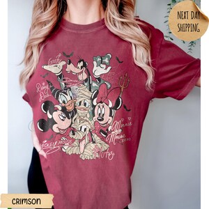 Peut inclure: T-shirt de couleur cramoisie avec un motif de personnages Disney en costumes d'Halloween. Mickey, Minnie, Donald, Dingo et Daisy Duck sont repr&eacute;sent&eacute;s. Le t-shirt porte les inscriptions manuscrites "Mickey Mouse" et "Minnie Mouse".