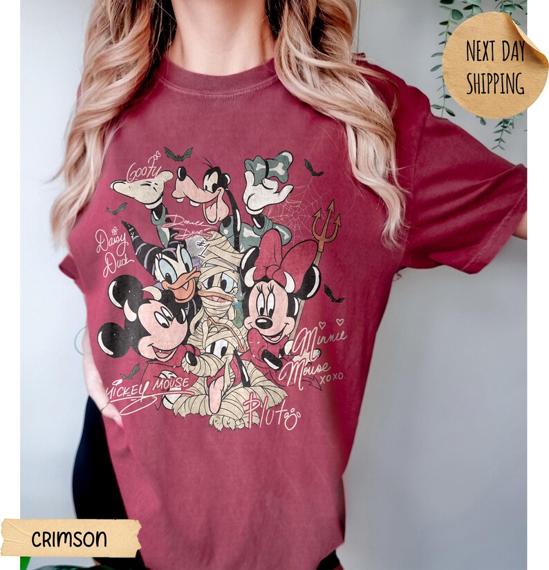 Peut inclure: T-shirt de couleur cramoisie avec un motif de personnages Disney en costumes d'Halloween. Mickey, Minnie, Donald, Dingo et Daisy Duck sont repr&eacute;sent&eacute;s. Le t-shirt porte les inscriptions manuscrites "Mickey Mouse" et "Minnie Mouse".