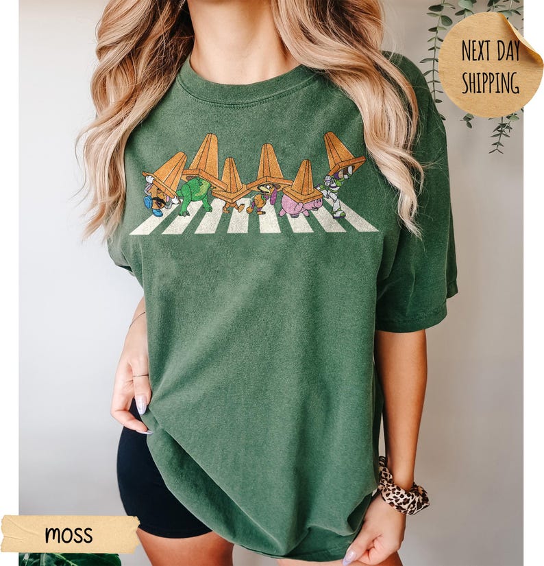 May include: A moss green short-sleeve t-shirt featuring a graphic of six cartoon characters walking in a line across a white crosswalk, each wearing an orange traffic cone on their head. The design is printed in a distressed, vintage-inspired style.